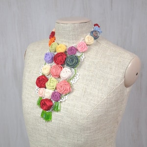 Pu&ograve; includere: Una collana floreale colorata realizzata con rose di stoffa con perline verdi che pendono dalla parte inferiore. La collana &egrave; esposta su un manichino rivestito in tessuto.