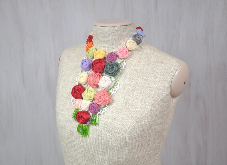 Pu&ograve; includere: Una collana floreale colorata realizzata con rose di stoffa con perline verdi che pendono dalla parte inferiore. La collana &egrave; esposta su un manichino rivestito in tessuto.