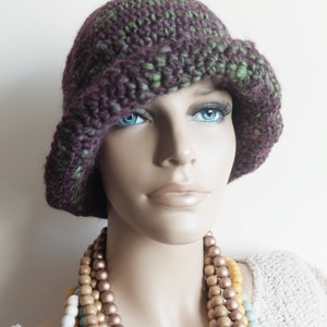 May include: A purple and green crocheted cloche hat with a wide brim. The hat is worn by a mannequin with a necklace of wooden beads.