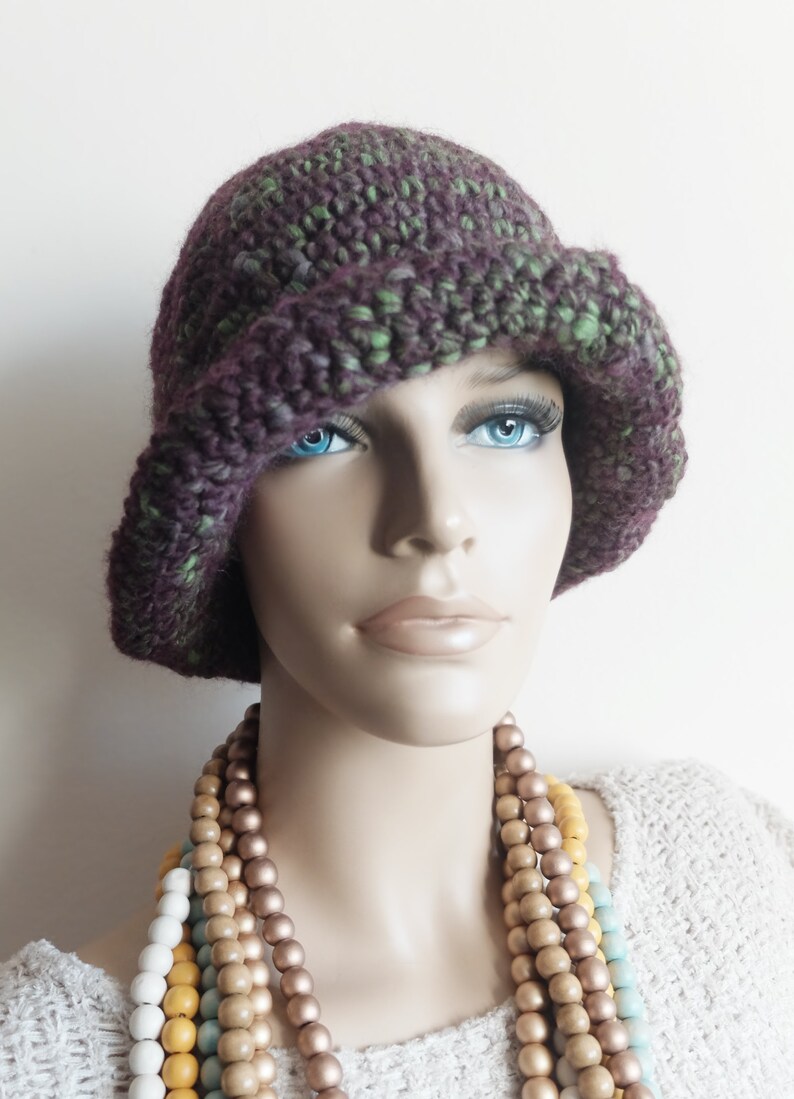 May include: A purple and green crocheted cloche hat with a wide brim. The hat is worn by a mannequin with a necklace of wooden beads.