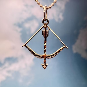 May include: A gold-coloured pendant necklace with a bow and arrow design. The arrow is positioned in the centre of the bow, with detailed fletching. The necklace chain is a delicate beaded style. The background is a soft blue sky with white clouds.