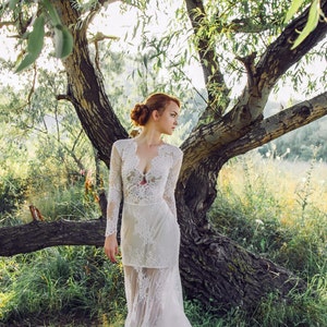 Puede incluir: Una mujer con un vestido de novia de encaje blanco se encuentra en un entorno de bosque. El vestido tiene un escote pronunciado y mangas largas. La mujer mira hacia un lado y su cabello est&aacute; recogido en un mo&ntilde;o suelto.