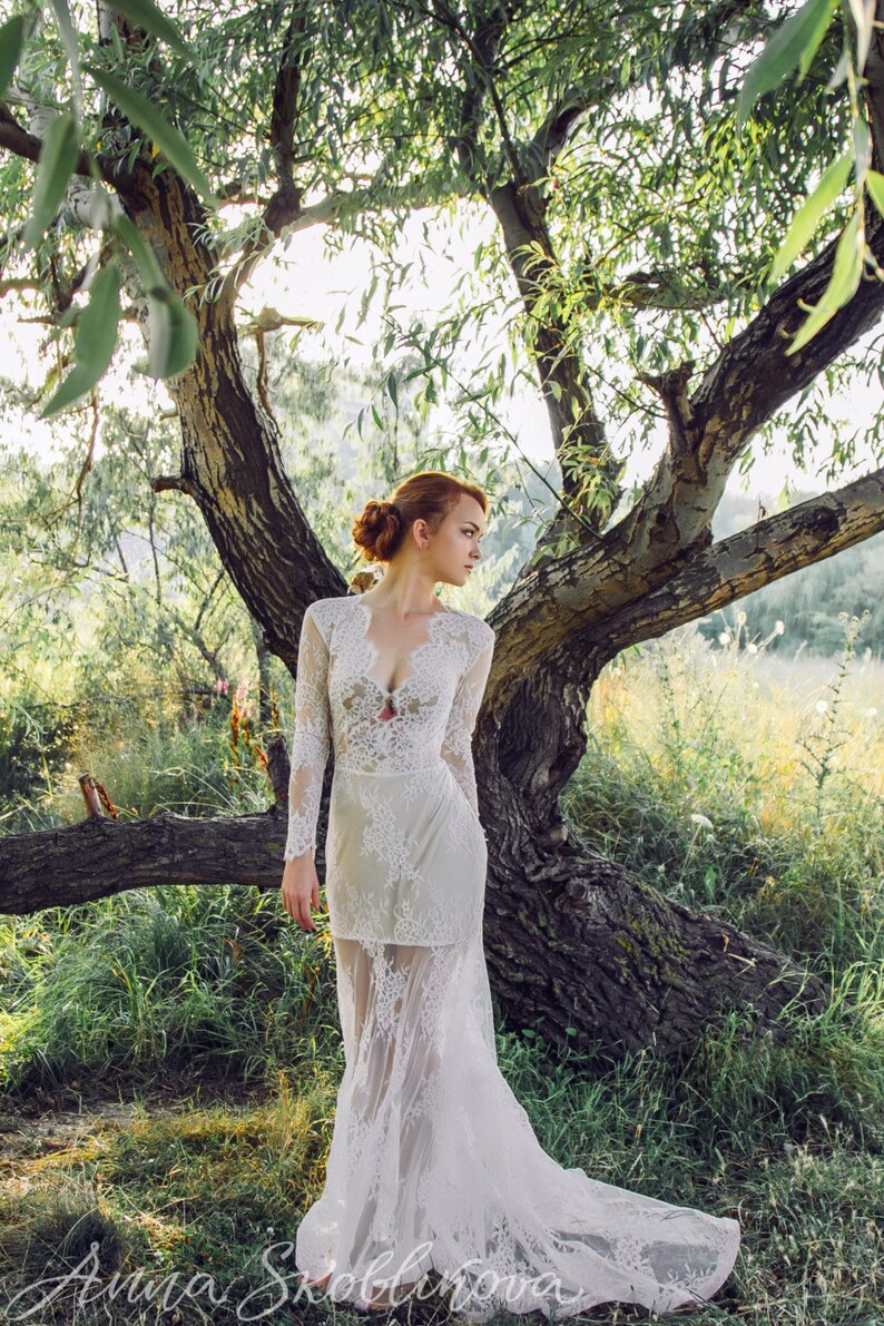 Puede incluir: Una mujer con un vestido de novia de encaje blanco se encuentra en un entorno de bosque. El vestido tiene un escote pronunciado y mangas largas. La mujer mira hacia un lado y su cabello est&aacute; recogido en un mo&ntilde;o suelto.