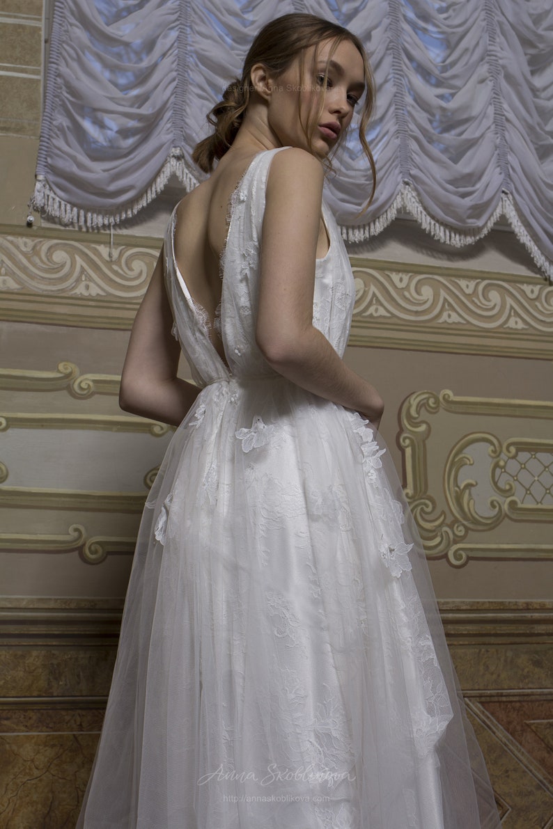 May include: A white wedding dress with a low back and a tulle skirt. The dress is decorated with delicate floral embroidery and has a flowing silhouette.