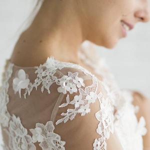 May include: Close-up of a white lace wedding dress with a sheer overlay. The lace features delicate floral patterns and is accented with small pearl-like embellishments.