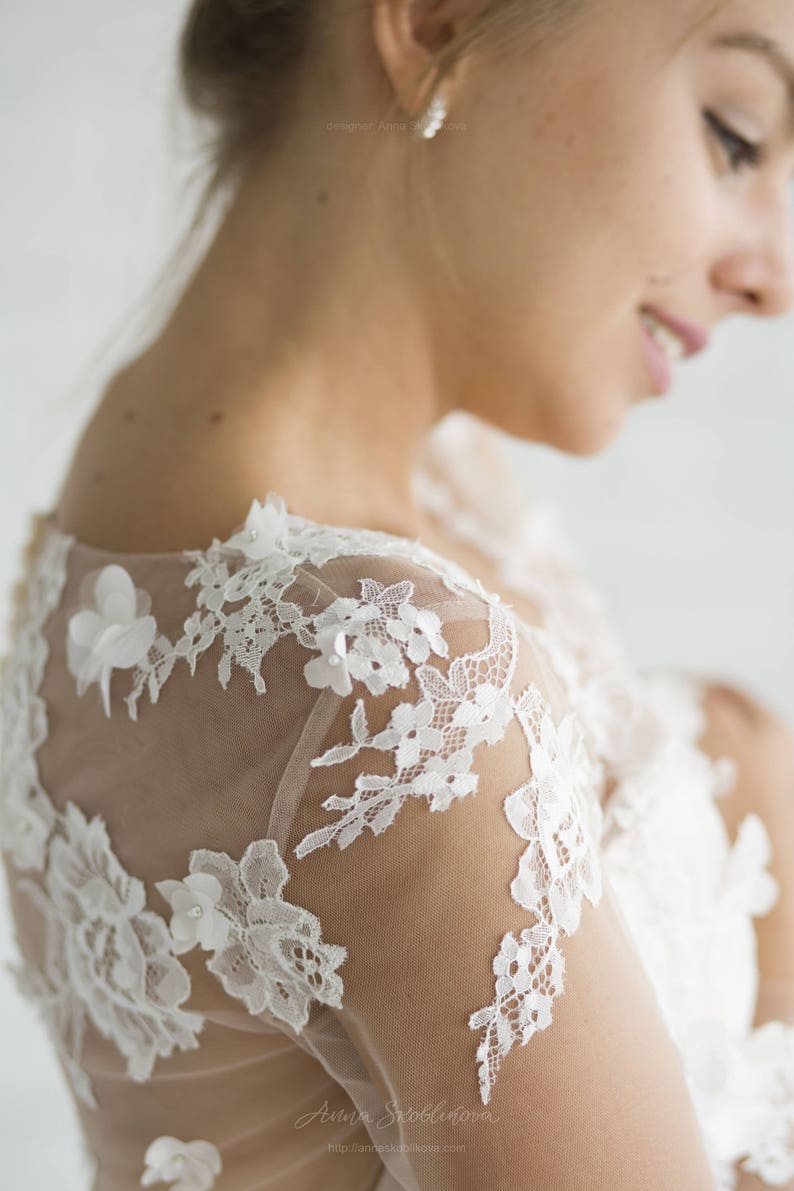 May include: Close-up of a white lace wedding dress with a sheer overlay. The lace features delicate floral patterns and is accented with small pearl-like embellishments.
