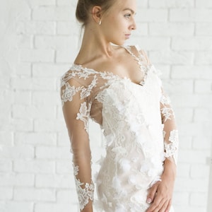 May include: A woman wearing a white lace wedding dress with a long sleeve design and floral embellishments. The dress has a fitted bodice and a flowing skirt.