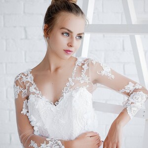 May include: A woman wearing a white lace wedding dress with a sweetheart neckline and long sleeves. She is standing in front of a white brick wall.