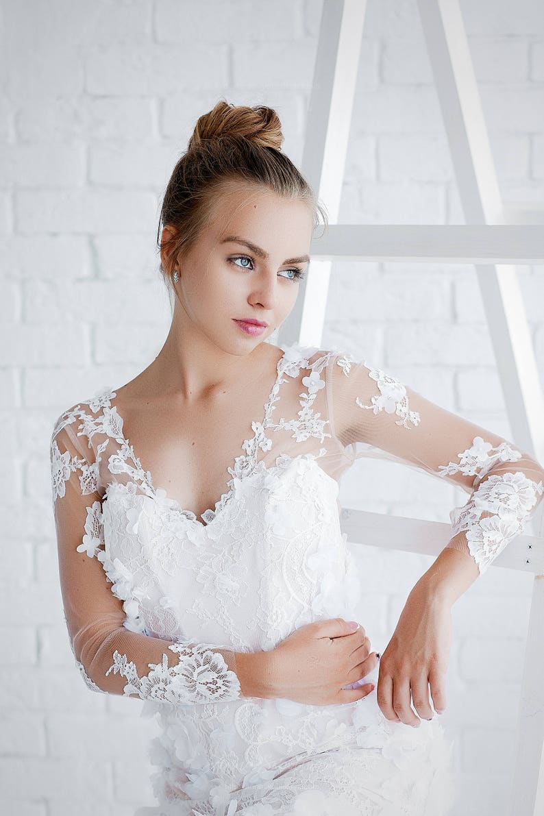May include: A woman wearing a white lace wedding dress with a sweetheart neckline and long sleeves. She is standing in front of a white brick wall.