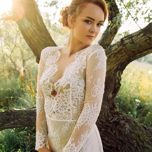 Puede incluir: Una mujer con un vestido blanco de encaje de manga larga. Est&aacute; de pie en un bosque con un &aacute;rbol detr&aacute;s de ella. El sol brilla a trav&eacute;s de las hojas del &aacute;rbol.