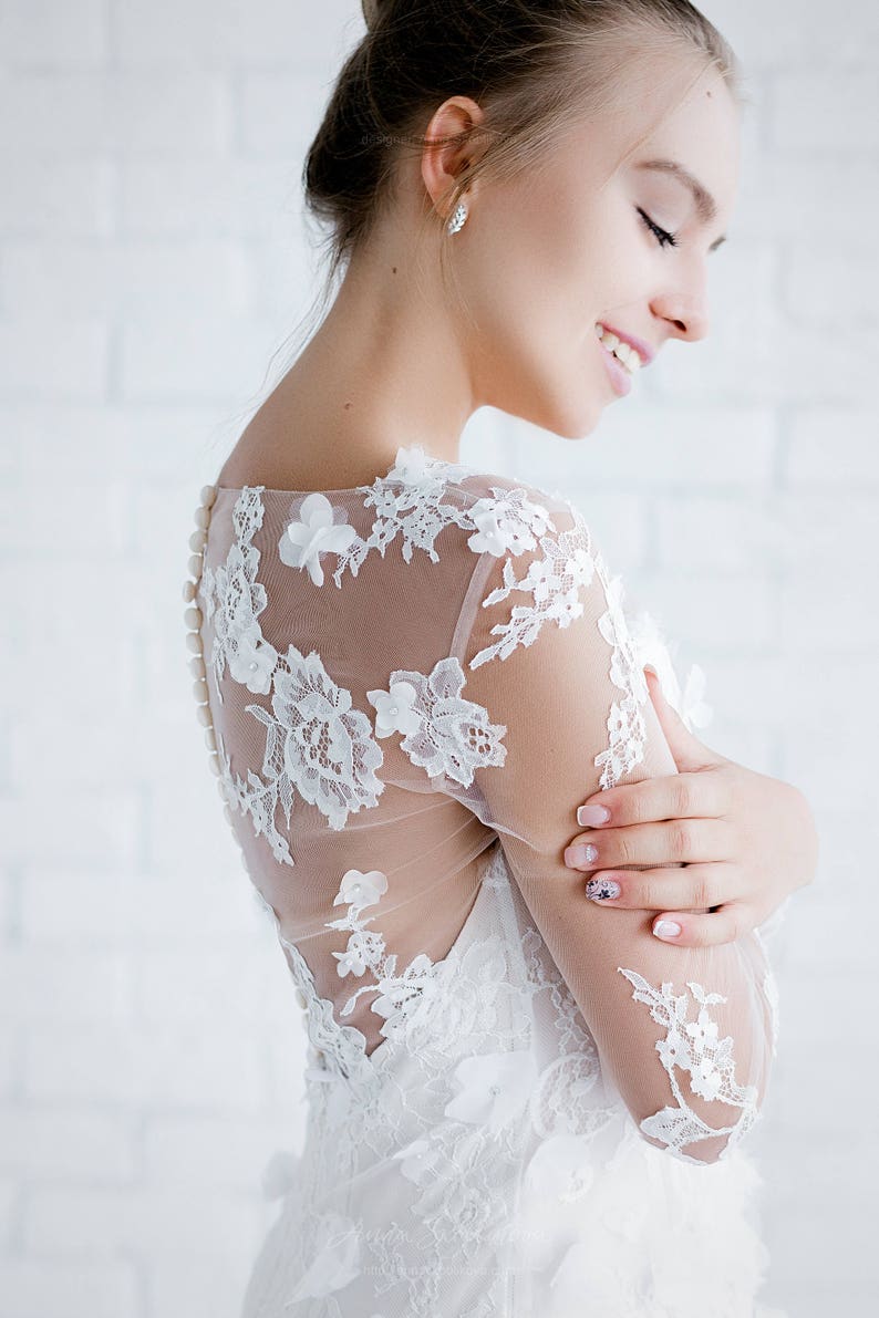 May include: A white lace wedding dress with a sheer bodice and long sleeves. The dress features delicate floral embroidery and a button closure at the back.