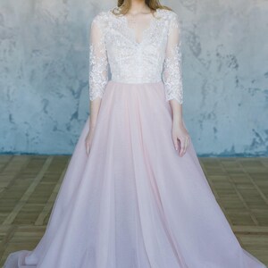 May include: A woman wearing a white lace top with a pink tulle skirt. The skirt is full and flowing, and the top has a V-neckline and three-quarter sleeves. The woman is standing in front of a grey wall.