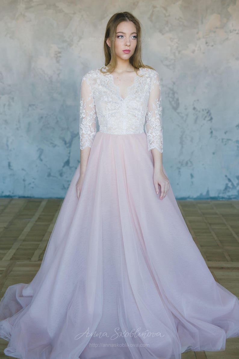 May include: A woman wearing a white lace top with a pink tulle skirt. The skirt is full and flowing, and the top has a V-neckline and three-quarter sleeves. The woman is standing in front of a grey wall.