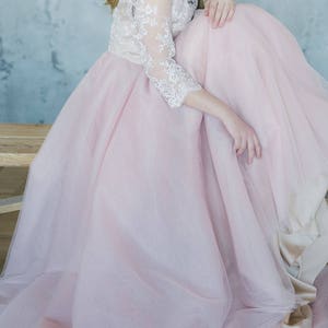 May include: A woman wearing a pink tulle skirt with a white lace top. The skirt is very full and the lace top has long sleeves. The woman is sitting on a wooden bench with her legs crossed.