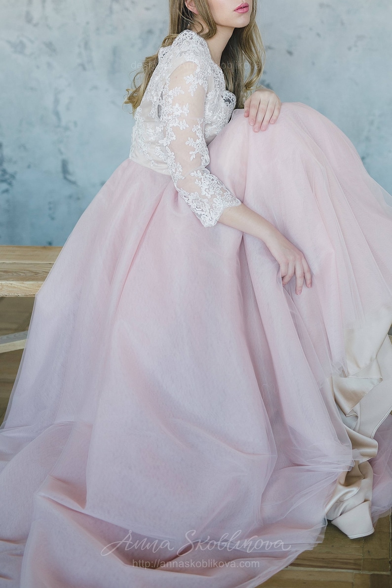 May include: A woman wearing a pink tulle skirt with a white lace top. The skirt is very full and the lace top has long sleeves. The woman is sitting on a wooden bench with her legs crossed.