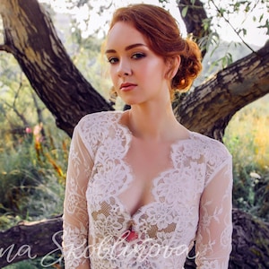 Puede incluir: Una mujer con cabello rojo lleva una blusa blanca de encaje con un escote en V profundo. Est&aacute; de pie en un campo con un &aacute;rbol detr&aacute;s de ella.