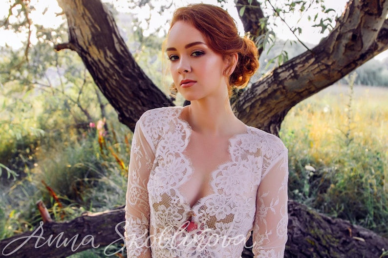 Puede incluir: Una mujer con cabello rojo lleva una blusa blanca de encaje con un escote en V profundo. Est&aacute; de pie en un campo con un &aacute;rbol detr&aacute;s de ella.