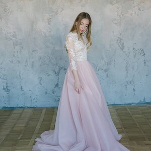 May include: A woman wearing a white lace top with long sleeves and a pink tulle skirt. The skirt is flowing and has a lot of volume. The woman is standing in front of a textured wall.