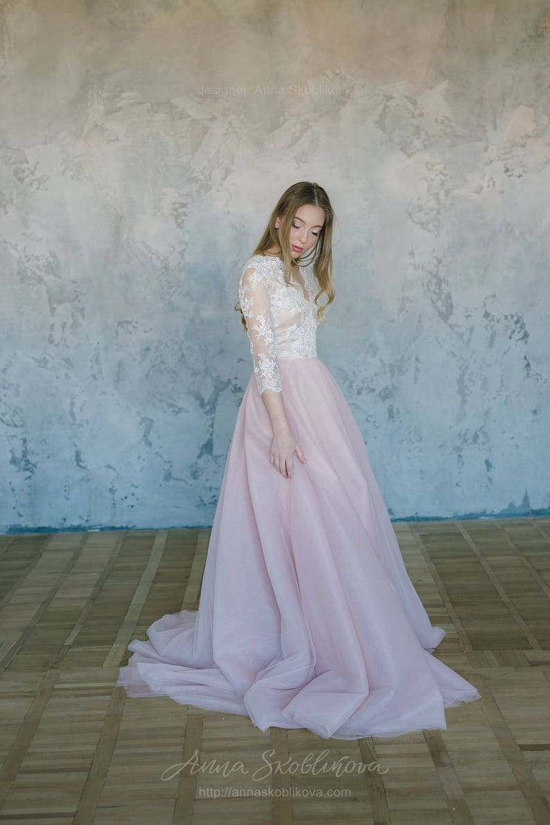 May include: A woman wearing a white lace top with long sleeves and a pink tulle skirt. The skirt is flowing and has a lot of volume. The woman is standing in front of a textured wall.