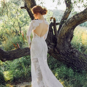 Puede incluir: Una mujer con un vestido de novia de encaje blanco con la espalda baja y mangas largas se encuentra en un entorno boscoso. El vestido tiene una cola fluida y una superposici&oacute;n transparente.