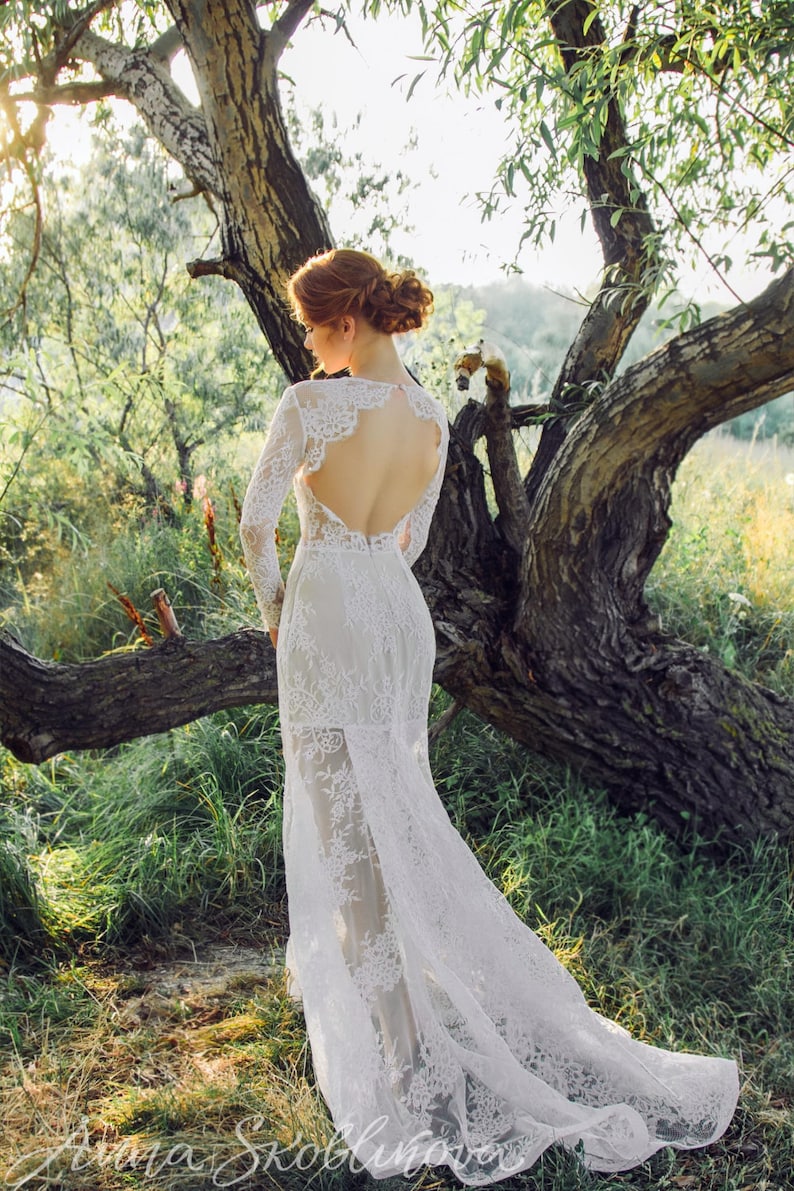 Puede incluir: Una mujer con un vestido de novia de encaje blanco con la espalda baja y mangas largas se encuentra en un entorno boscoso. El vestido tiene una cola fluida y una superposici&oacute;n transparente.