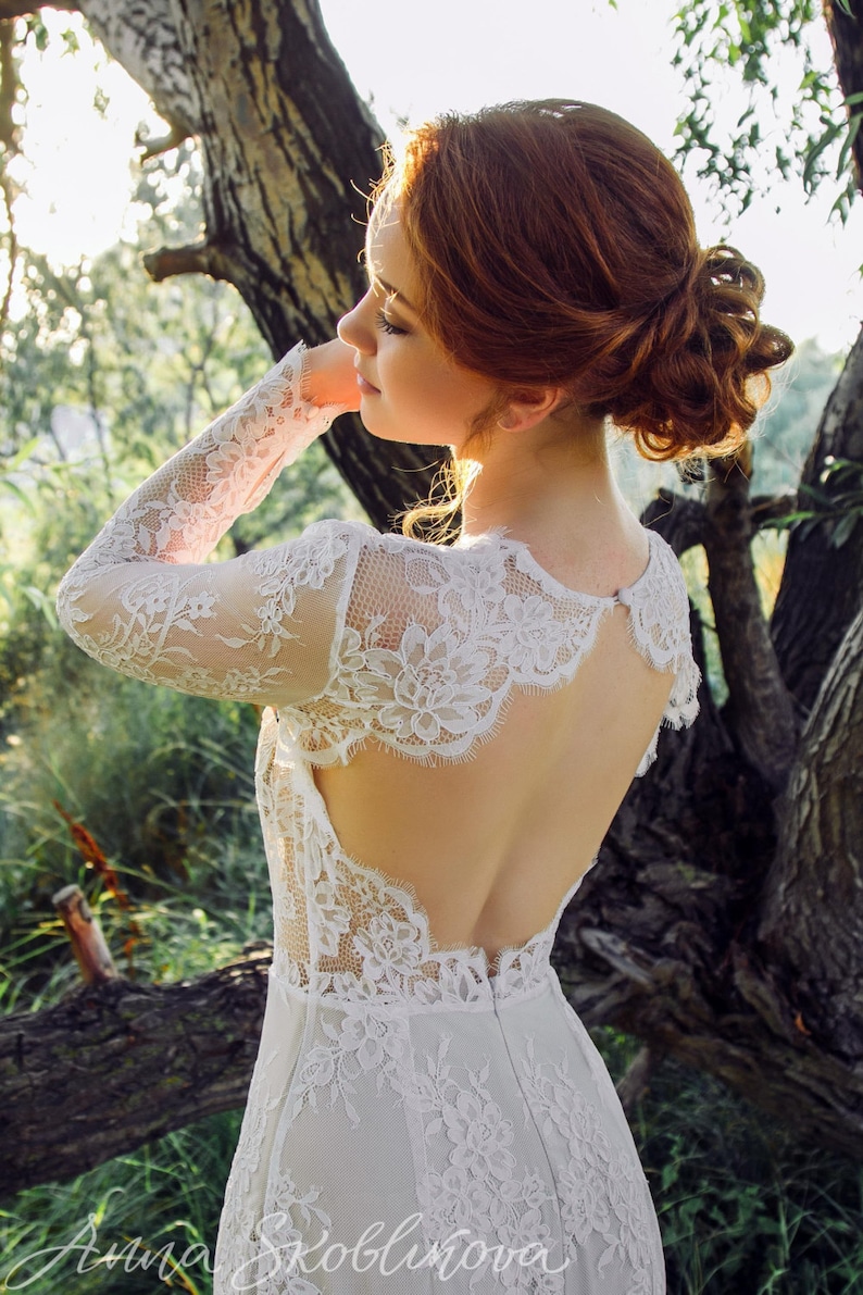Puede incluir: Una mujer con cabello rojo lleva un vestido de novia de encaje blanco con la espalda baja y mangas largas. El vestido tiene un delicado patr&oacute;n floral y un escote festoneado.