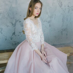 May include: A woman wearing a long, pink tulle dress with a white lace bodice. She is sitting on a wooden bench with a neutral coloured wall in the background.