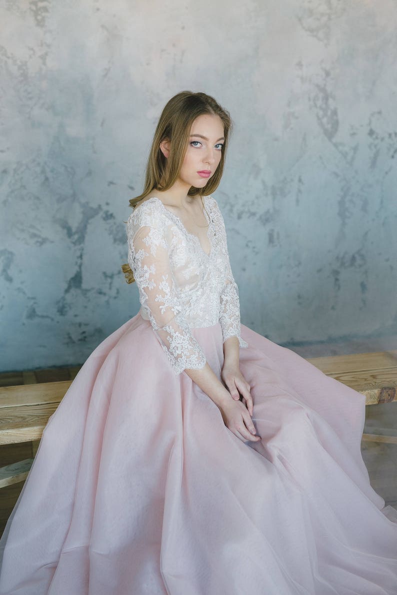 May include: A woman wearing a long, pink tulle dress with a white lace bodice. She is sitting on a wooden bench with a neutral coloured wall in the background.