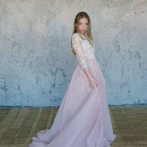 May include: A woman wearing a white lace top and a long, flowing pink tulle skirt. The skirt is gathered at the waist and has a slight train. The woman is standing in front of a textured wall.