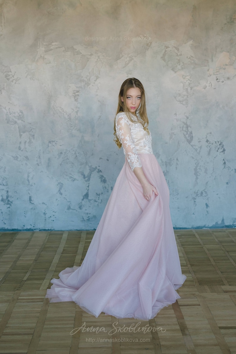 May include: A woman wearing a white lace top and a long, flowing pink tulle skirt. The skirt is gathered at the waist and has a slight train. The woman is standing in front of a textured wall.