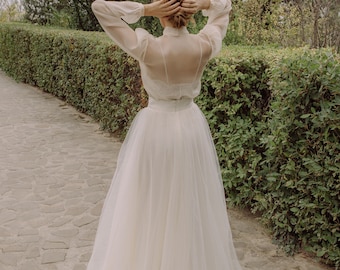 May include: A woman wearing a white sheer blouse with a tulle skirt. The skirt is long and flowing, and the blouse has a lace design. The woman is standing on a stone path in a garden.