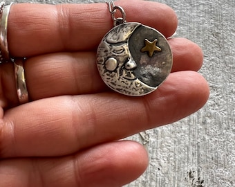 Puede incluir: Un collar de plata con un colgante de media luna con una cara y una estrella dorada. El colgante es texturizado y tiene un aspecto rústico.