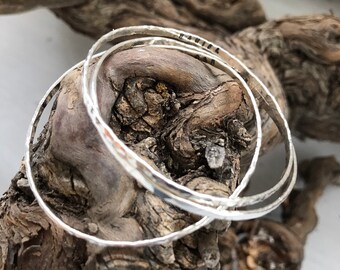 May include: Three silver bangles stacked on top of each other. The bangles are hammered and have a textured surface. The top bangle has the word "PAIGE" inscribed on it.