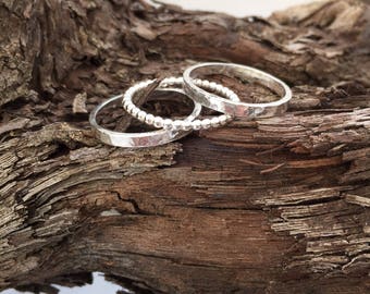 May include: Three silver rings with varied textures are displayed on a piece of wood. One ring has a hammered finish, another features small beaded accents, and the third has a smooth, flat surface. The rings are set against a backdrop of brown wood.