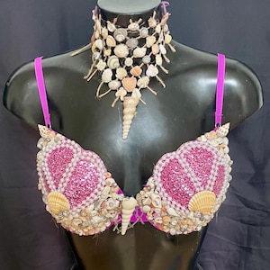 Könnte beinhalten: Ein BH- und Choker-Set zum Thema Meerjungfrau. Der BH ist pink mit Glitzer und Muscheln. Der Choker besteht aus Muscheln und Bindfäden. Das Set wird auf einer schwarzen Schaufensterpuppe präsentiert.