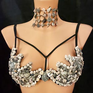 May include: A seashell bra and choker set on a mannequin. The bra has a black strappy design, adorned with white, grey, and beige seashells. The choker is a grid of seashells. The background is black.