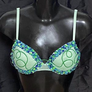 May include: A light green bra with blue and green embellishments. The bra features a decorative design on the cups and is displayed on a black mannequin. The bra straps are a matching light green color.