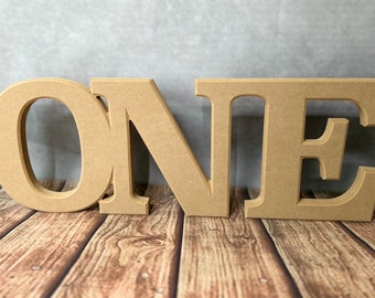 May include: Wooden letters spelling out the word "ONE" in a light brown color. The letters are standing on a wooden surface.