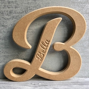 May include: A wooden letter B with a cursive font. The letter is unfinished and ready to be painted. The name "Bella" is engraved on the letter.