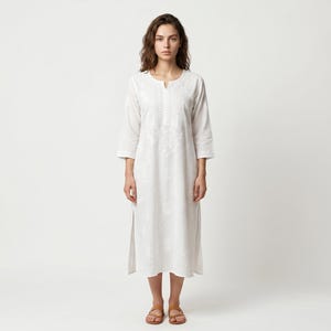 May include: A white, embroidered midi dress with three-quarter sleeves and a split hem. The dress features intricate floral embroidery around the neckline and down the front. The model is wearing brown sandals.