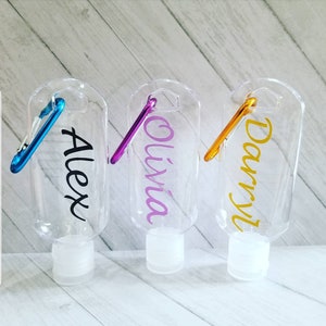 May include: Three clear plastic hand sanitizer bottles with a carabiner clip. The bottles are labeled with the names "Alex", "Olivia", and "Darryl".