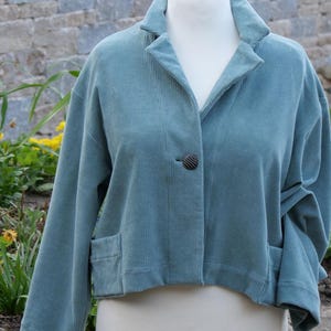 Könnte beinhalten: Eine tealfarbene Cordjacke mit Reverskragen, zwei Vordertaschen und einem einzelnen dunklen Knopfverschluss. Die Jacke wird auf einer weißen Schaufensterpuppe präsentiert und hat eine verkürzte Passform. Der Stoff hat eine vertikale Rippenstruktur.