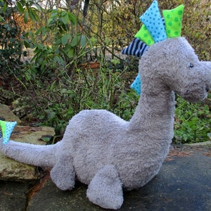 Könnte beinhalten: Ein graues Plüsch-Dinosaurierspielzeug mit einem blauen, grünen und schwarzen gemusterten Kamm. Der Dinosaurier hat einen langen Schwanz und ein freundliches Gesicht.