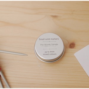 May include: A small tin of mixed color stitch markers for knitting or crocheting. The tin is labeled "Small solid markers" and "The Woolly Tansle" with the text "up to 4mm mixed colours".