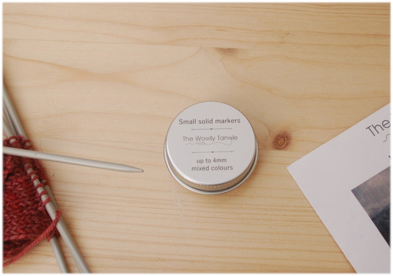May include: A small tin of mixed color stitch markers for knitting or crocheting. The tin is labeled "Small solid markers" and "The Woolly Tansle" with the text "up to 4mm mixed colours".