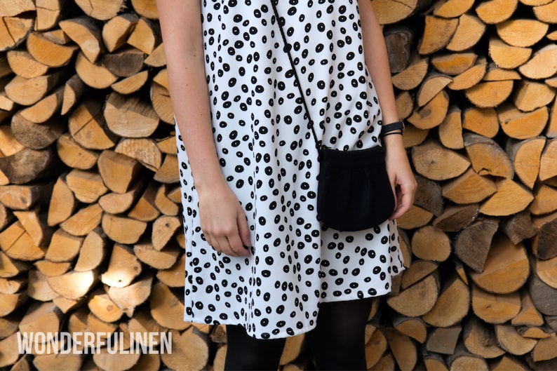 Pu&ograve; includere: Un vestito bianco con un motivo a pois neri e una borsa a tracolla nera. Il vestito &egrave; indossato da una persona in piedi davanti a un muro di legna da ardere impilata.
