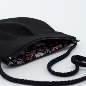 Pu&ograve; includere: Una borsa a tracolla nera con chiusura a zip e fodera con motivo floreale. La borsa ha una lunga tracolla in corda nera.