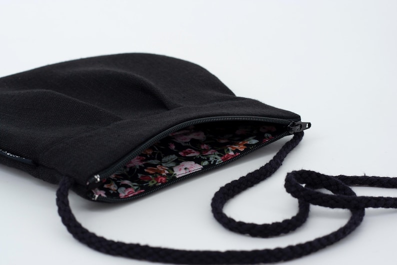 Pu&ograve; includere: Una borsa a tracolla nera con chiusura a zip e fodera con motivo floreale. La borsa ha una lunga tracolla in corda nera.