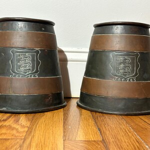 May include: Two antique metal tankards with a dark gray and copper color scheme. Each tankard features a decorative emblem and the word "Jersey" etched on the front. The tankards are displayed on a wooden surface.