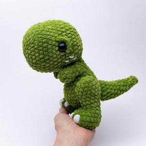 Puede incluir: Un juguete de dinosaurio verde tejido a ganchillo con una cabeza grande, brazos pequeños y una cola. El dinosaurio tiene ojos negros y detalles blancos en los pies y la boca. El juguete está hecho de un material afelpado texturizado.